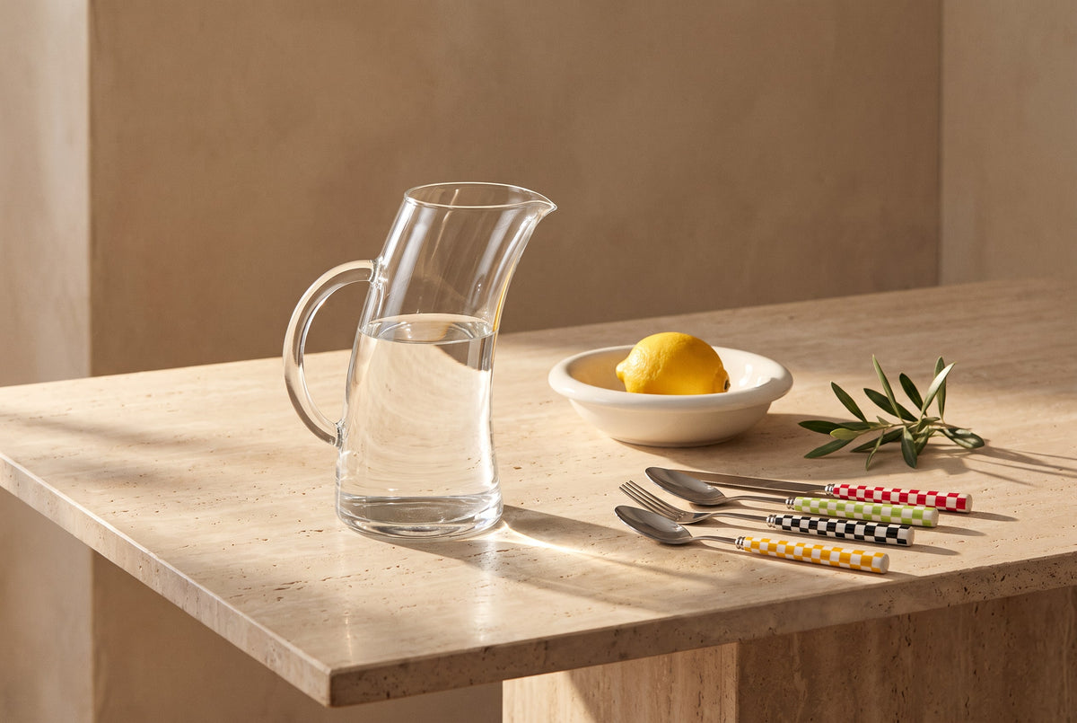 Glass water jug, white ceramic bowl of lemons and checkered flatware on a travertine table