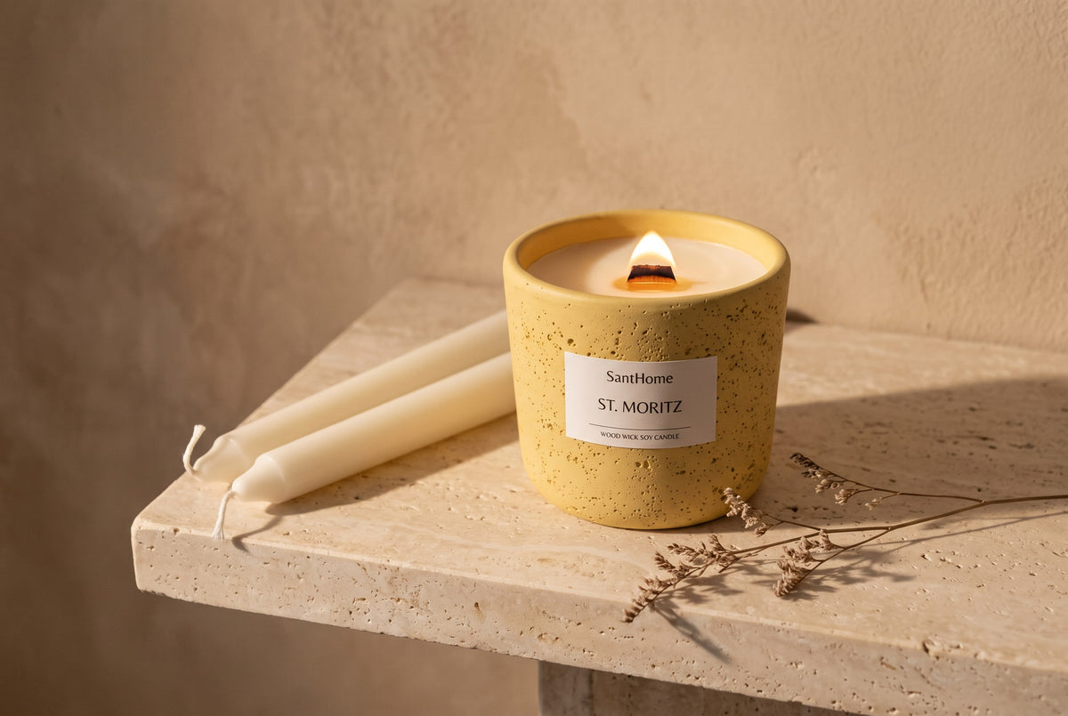 Lit wood wick soy candle in a yellow ceramic pot beside ivory taper candles on stone