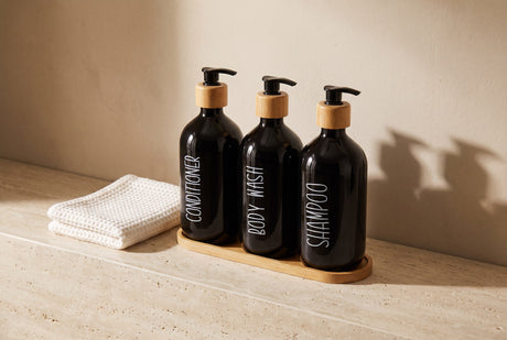 Black glass bathroom bottle trio labelled conditioner, body wash and shampoo beside a folded towel