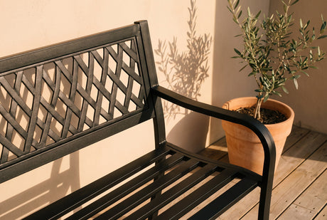Black cast-metal garden bench with diamond lattice backrest beside a potted olive tree