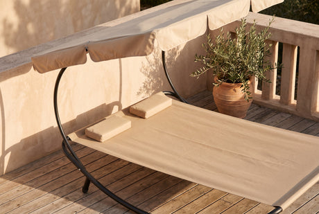 Two-person sun lounger with beige canopy and bolster pillows beside a potted olive branch