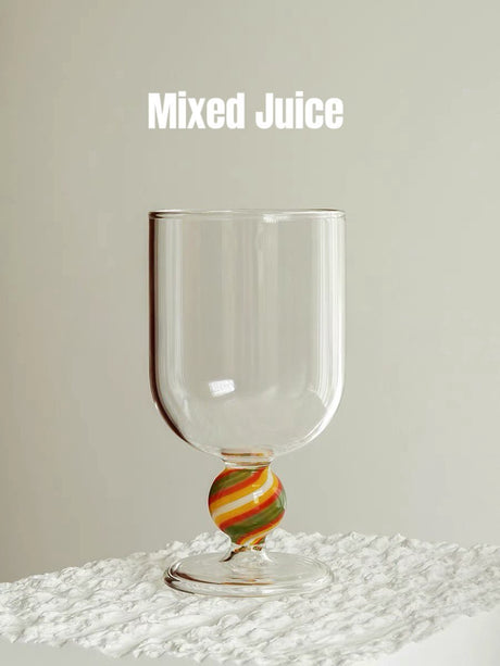 A clear wine glass with a striped orange, yellow and green gumball stem, on a textured white surface
