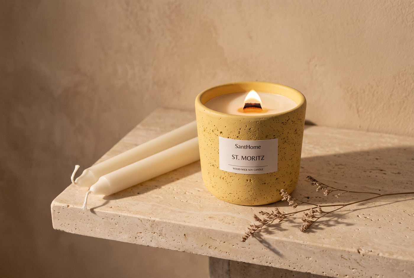 Lit wood wick soy candle in a yellow ceramic pot beside ivory taper candles on stone